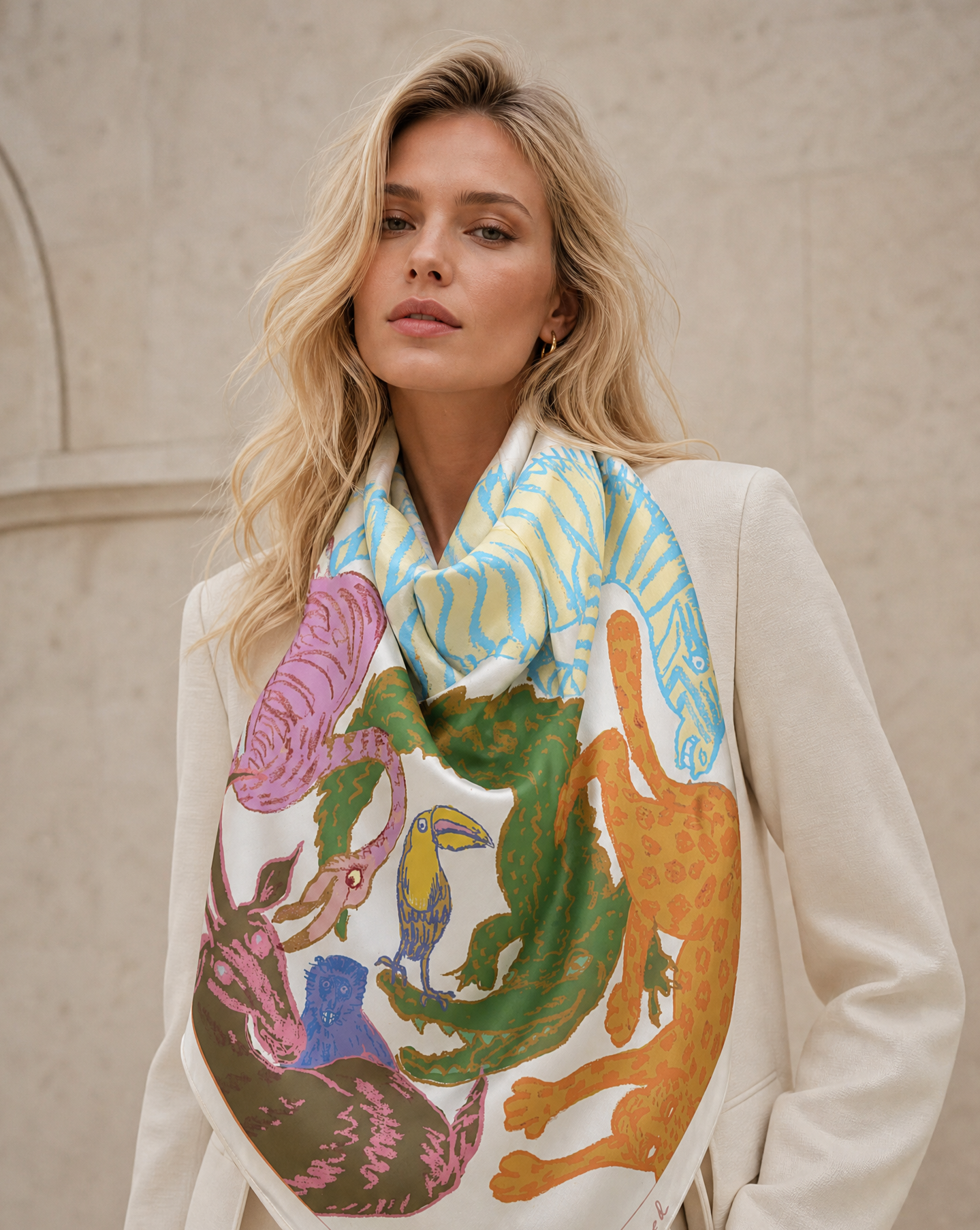 A blonde woman wearing a cream-colored blazer and a large, vibrant square scarf with a multicolor animal and bird illustration, posed against a light beige stone wall.